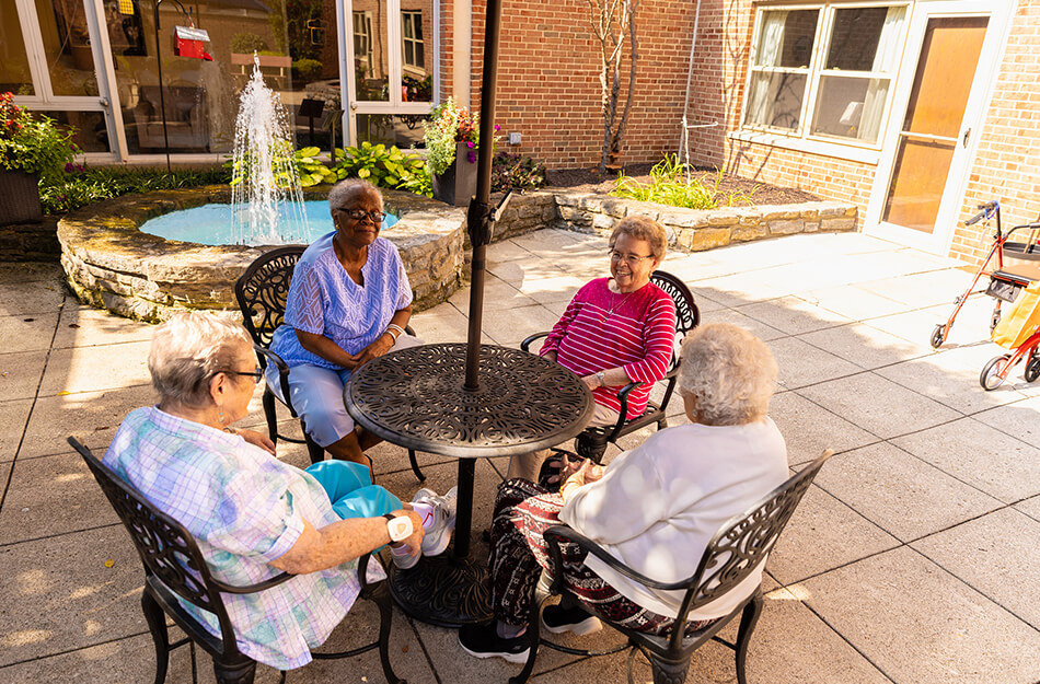 Four elderly women sitting around a table near a fountain in a courtyard.
