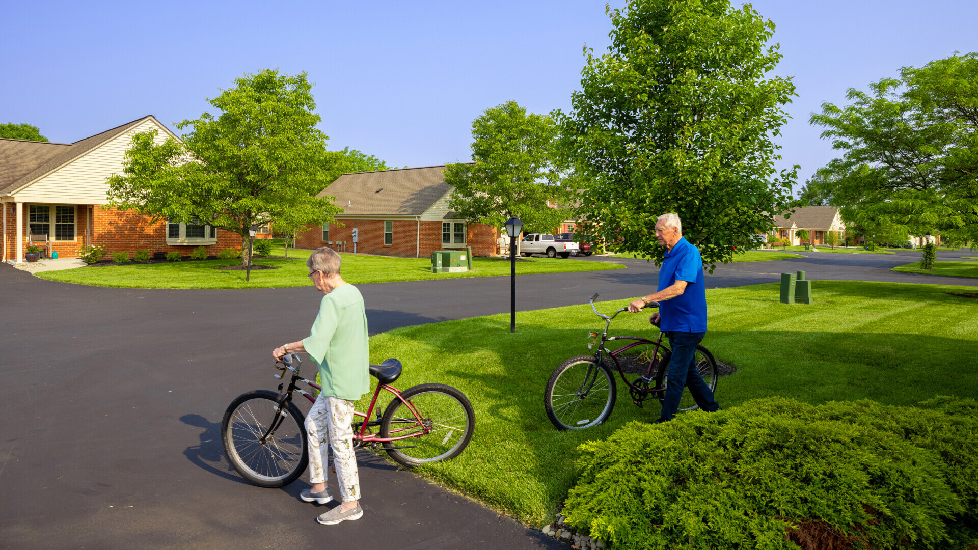 Two residents wheel their bikes out into a summer day in a vibrant retirement community.