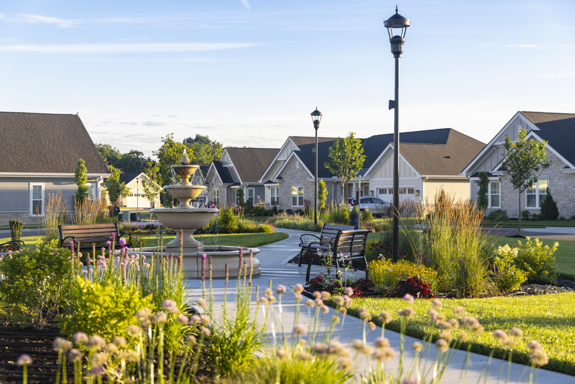 Exterior shot of a well-maintained community park at dusk. A multi-tiered stone fountain sits centrally on a paved walkway lined with benches and lush landscaping including ornamental grasses and flowering plants. Traditional style homes with dark brown roofs are visible in the background under a clear sky. The scene evokes a sense of peace and upscale residential living at the Christian Village at Mason.