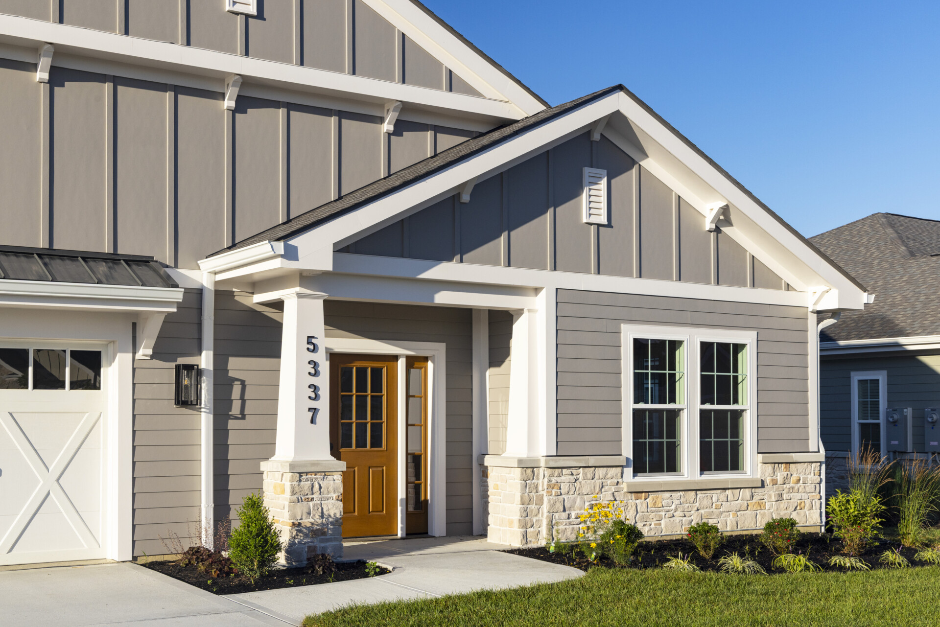 Exterior view of a modern farmhouse with gray vertical board and batten siding, a bright yellow front door framed by stone pillars, and green window frames. A garage door with X-shaped detailing is visible to the left. The scene is bathed in sunlight under a clear blue sky at the Christian Village at Mason.