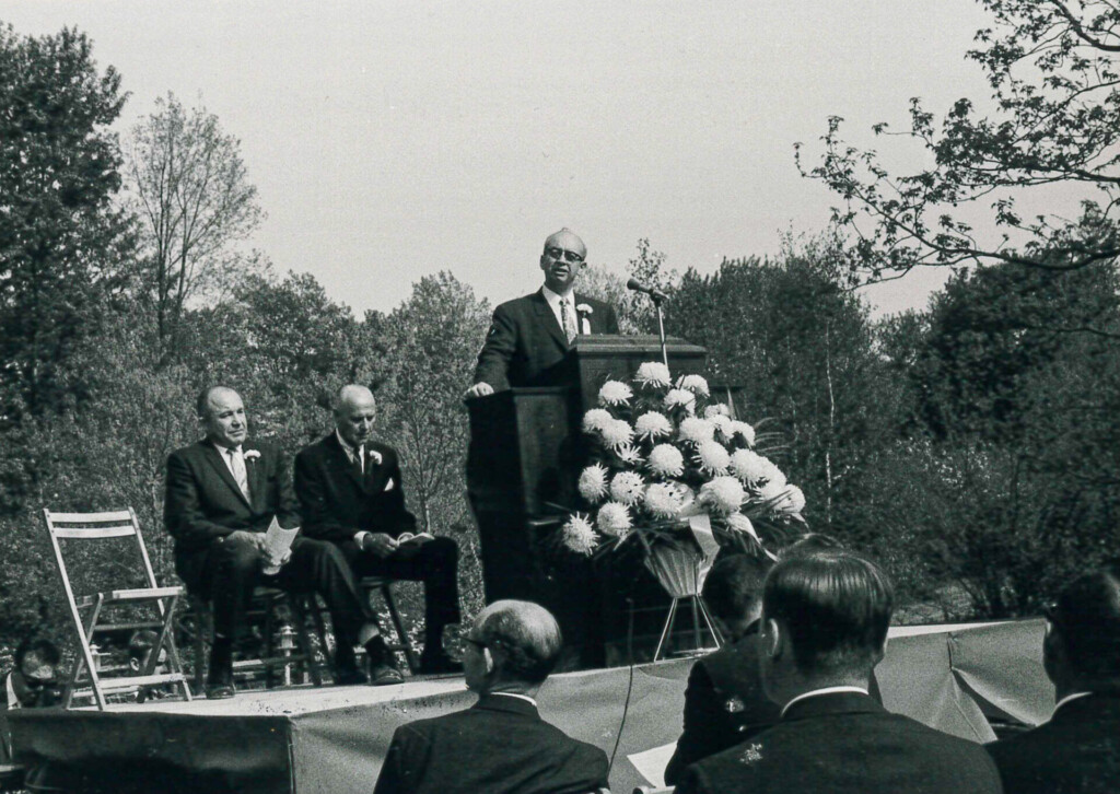 Suited man at podium on an outdoor stage speaks to a small crowd on a sunny day in May 1966. The photo is old and grainy, in black and white.