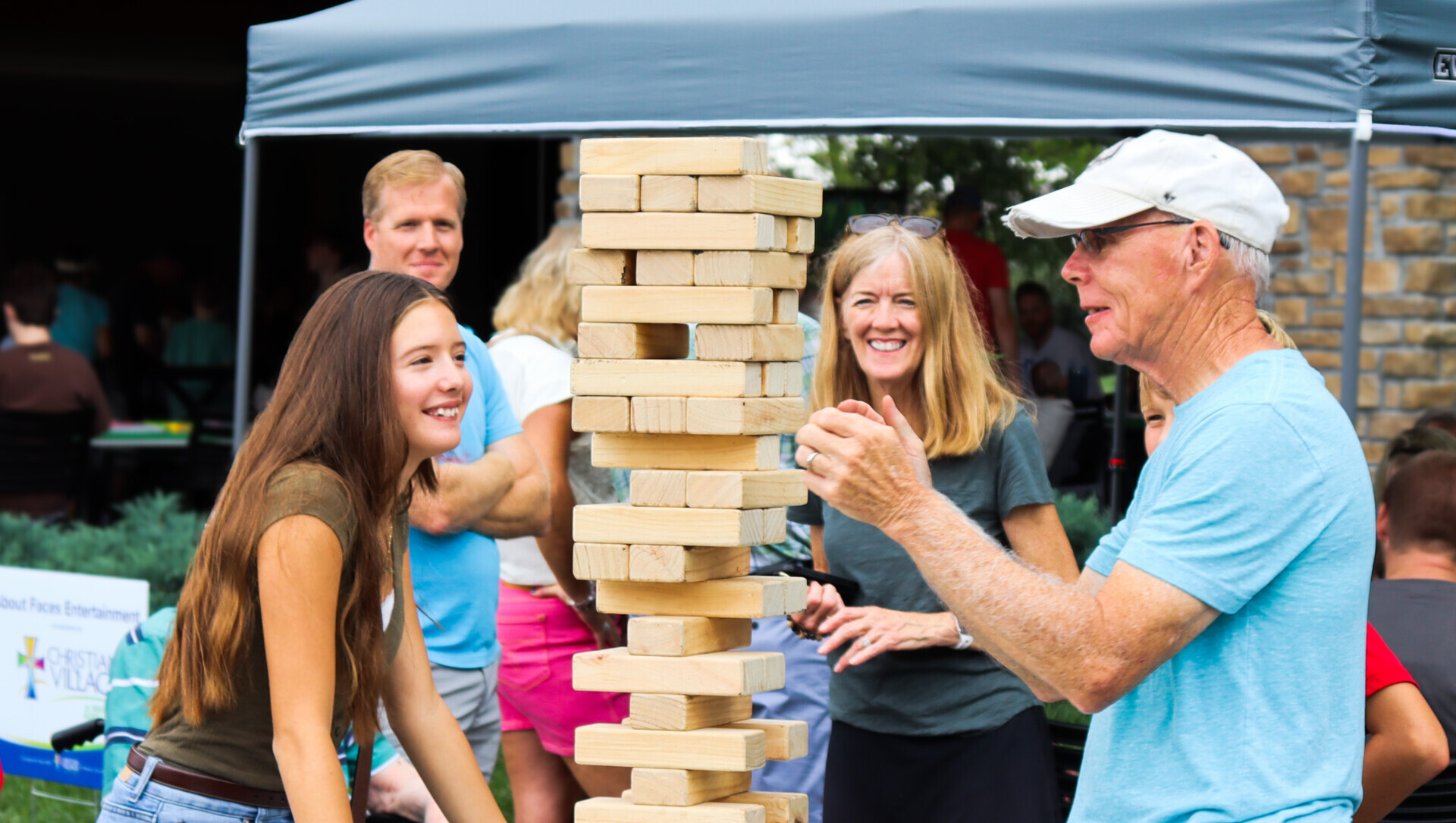 A grandfather plays jumbo jenga outside with his granddaughter as their family stands around the table watching.